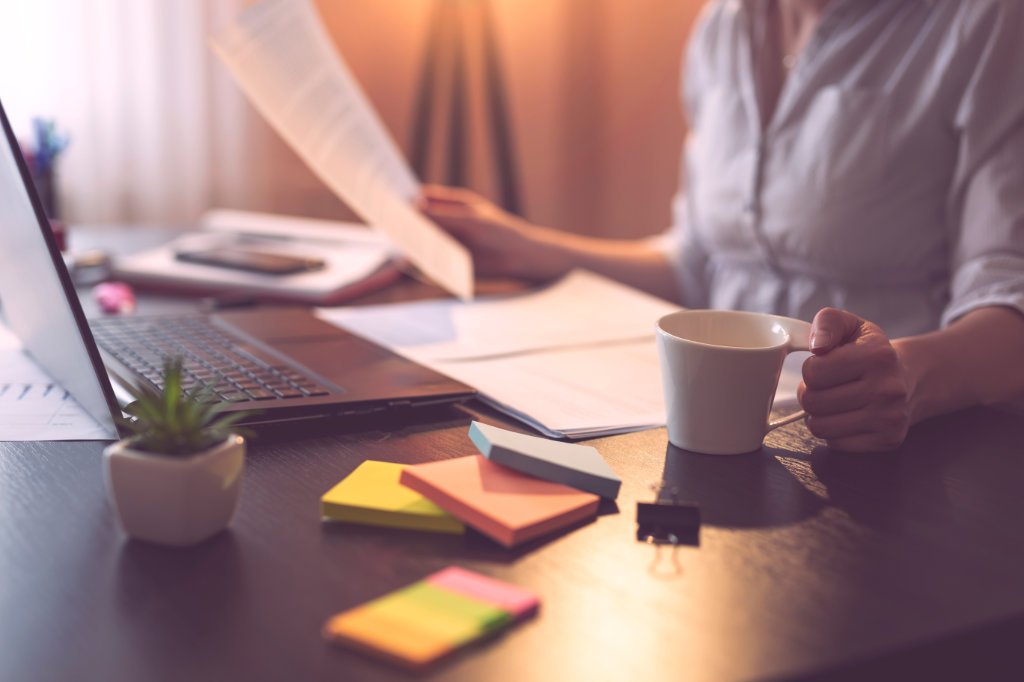 Businesswoman sitting at her desk in a home office, reviewing a contract and drinking coffee. Selective focus on the cup and the hand